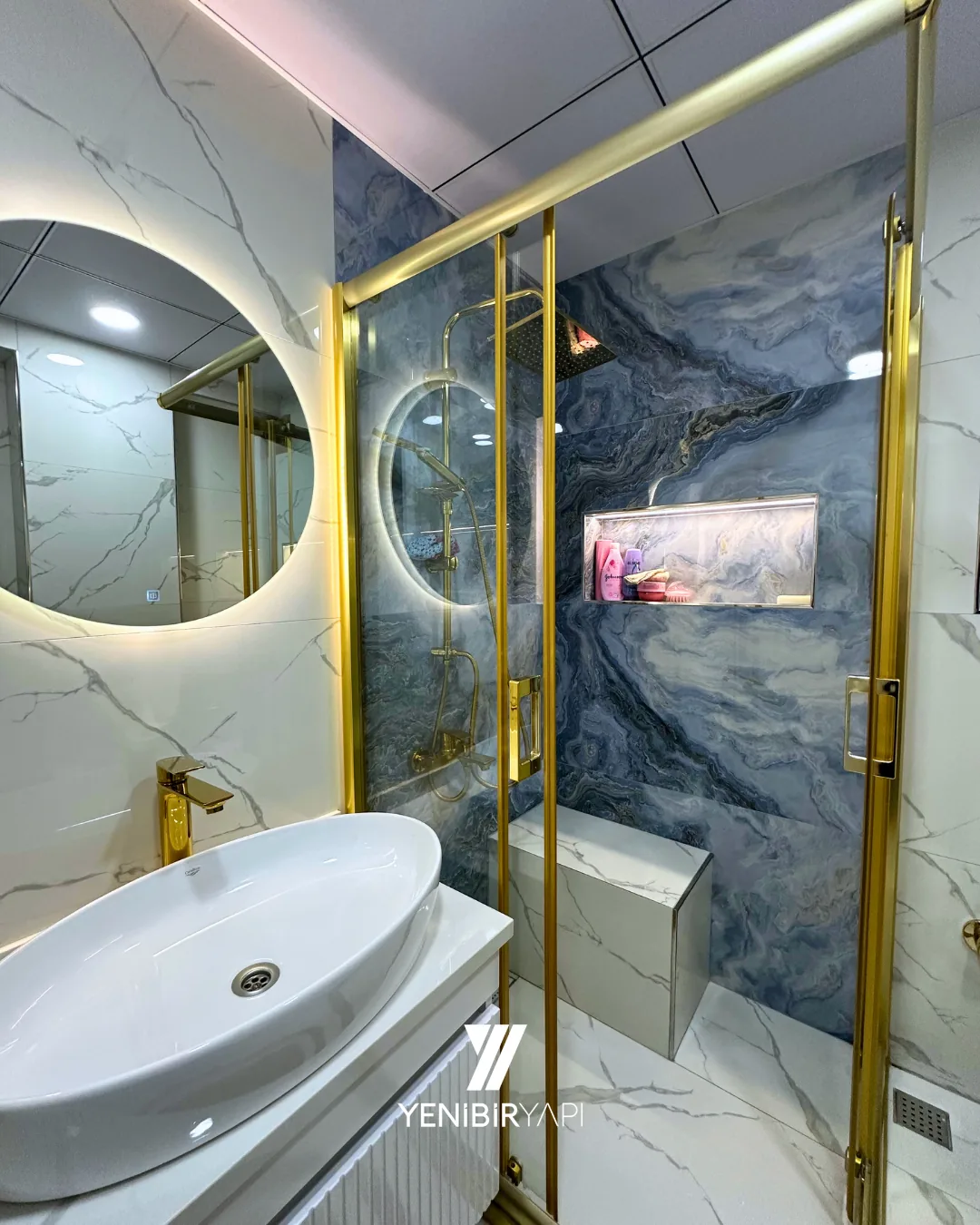 Tamamlanmış banyo: mavi damarlı mermer duvar, altın rengi çerçeveli duşakabin ve arkadan aydınlatmalı yuvarlak ayna